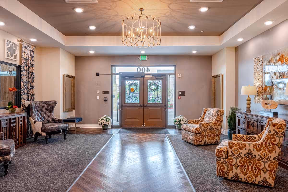 Bright furnished senior living lobby with patterned armchairs, a chandelier, and double entrance doors.