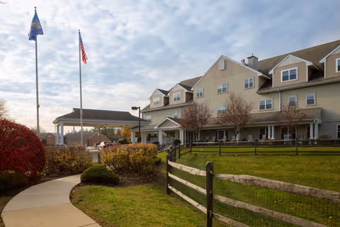 Exterior view of Simpson Meadows Retirement Community showing a multi-story building with beige siding, a covered entrance with an American flag and another flag on poles, a curved sidewalk, green lawn, bushes, and a wooden fence under a partly cloudy sky.
