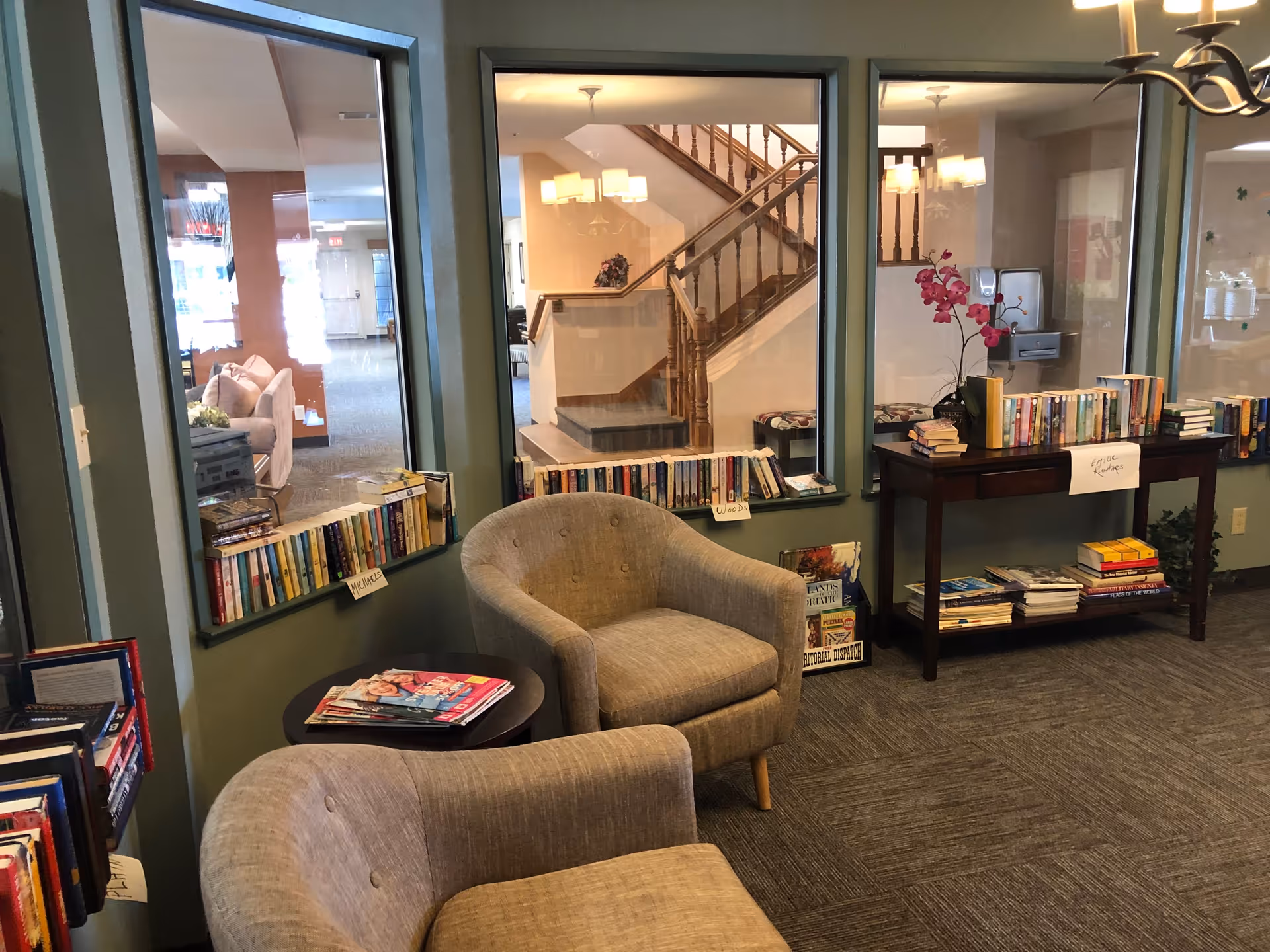 Cozy reading nook in a retirement community with two beige armchairs, a small round table with magazines, and bookshelves filled with books. Large windows show a staircase and other seating areas in the background.