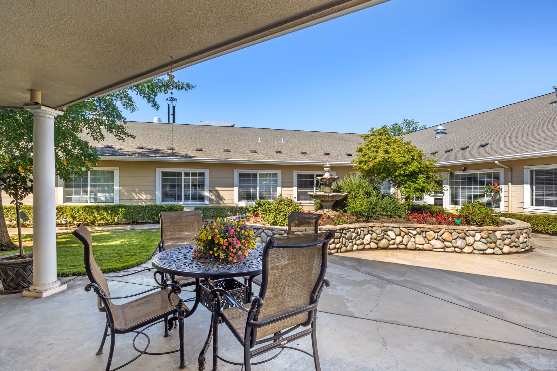 Outdoor patio area with a round metal table and four chairs. The table has a colorful flower arrangement in the center. Surrounding the patio is a garden with a stone wall, various plants, bushes, and a multi-tiered water fountain. The building with beige siding and white-trimmed windows is visible in the background under a clear blue sky.