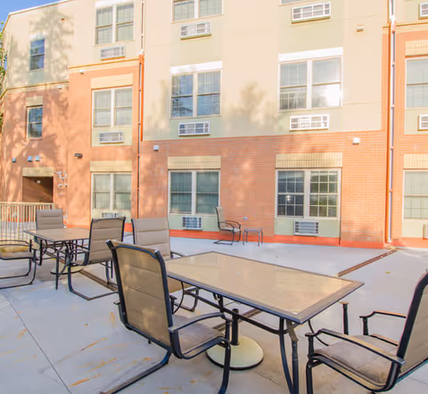 Outdoor patio area with several glass-top tables and cushioned chairs set on a concrete surface, adjacent to a multi-story brick and beige building with multiple windows and air conditioning units.