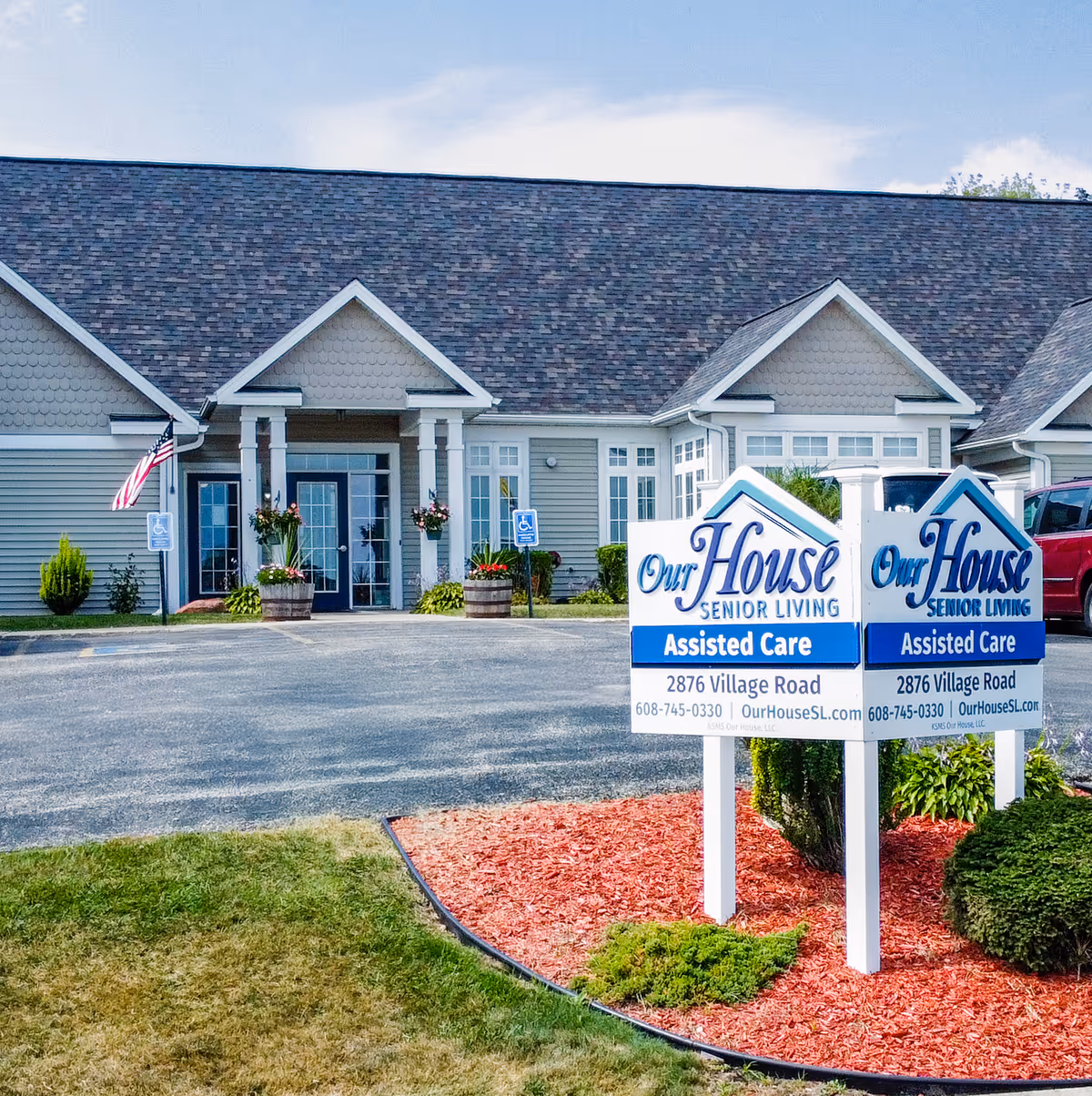 Exterior view of Our House Senior Living - Portage Assisted Care facility showing the front entrance with columns, windows, and an American flag. In front, there is a sign with the facility name, address, phone number, and website. The area around the sign is landscaped with grass, mulch, and bushes.