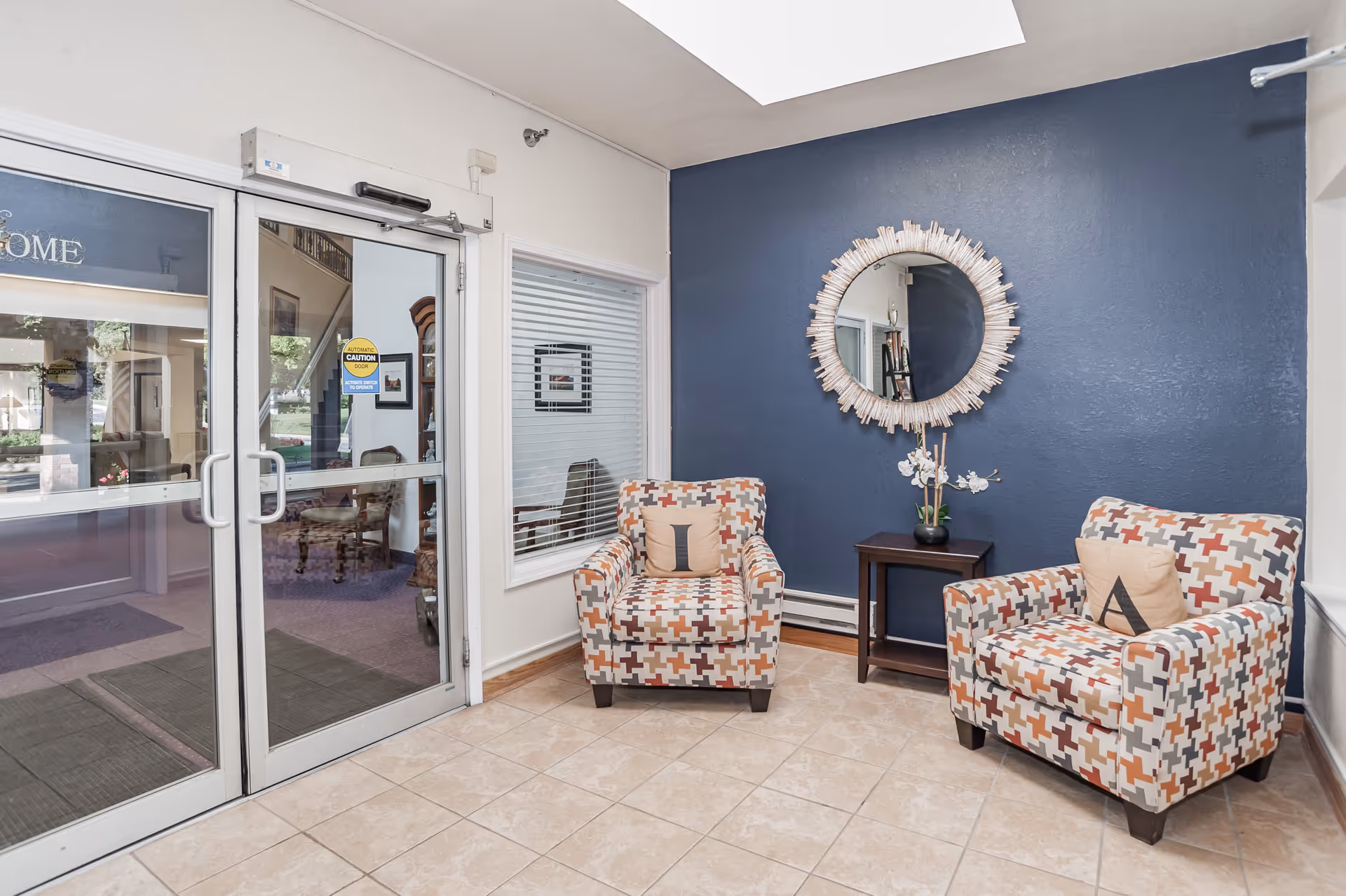 A small seating area near a glass entrance door with two patterned armchairs, each with a decorative pillow labeled 'I' and 'A'. A small wooden side table with a potted orchid is placed between the chairs. A round decorative mirror hangs on a dark blue accent wall behind the chairs. The floor is tiled and there is a window with blinds next to the door.