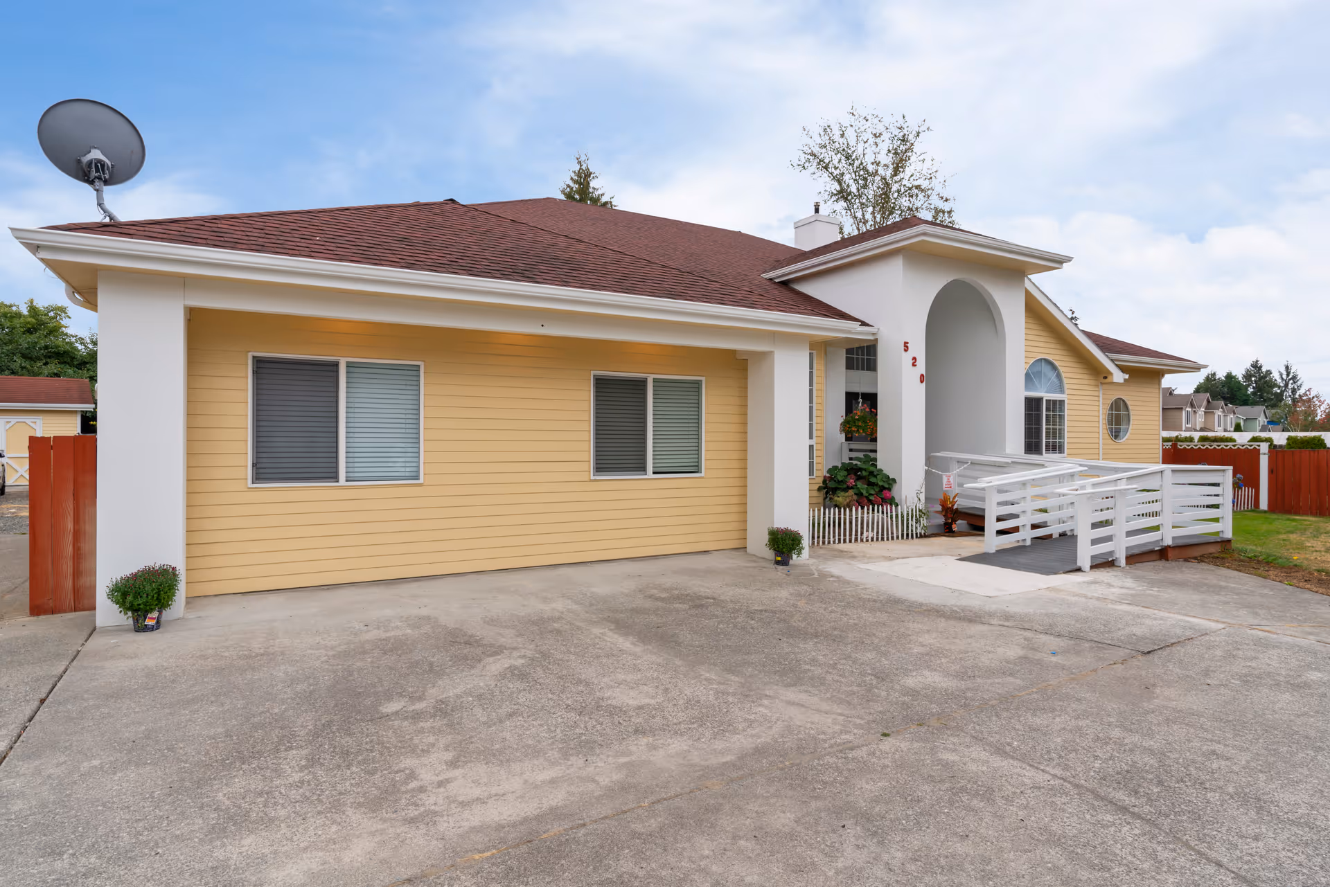 Front exterior of a single-story yellow house with a white wheelchair ramp, potted plants, and a concrete driveway.
