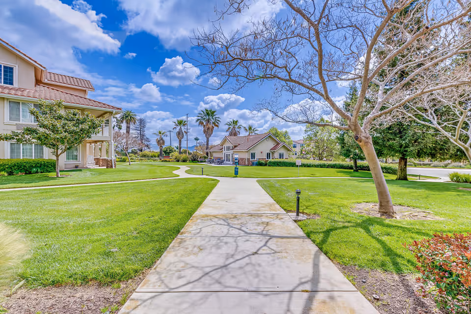 A paved walkway leads through a well-maintained green lawn with trees and shrubs on either side. To the left is a multi-story building with a tiled roof and large windows. In the background, there is a smaller building with a similar roof style. Palm trees and a partly cloudy blue sky are visible in the distance.