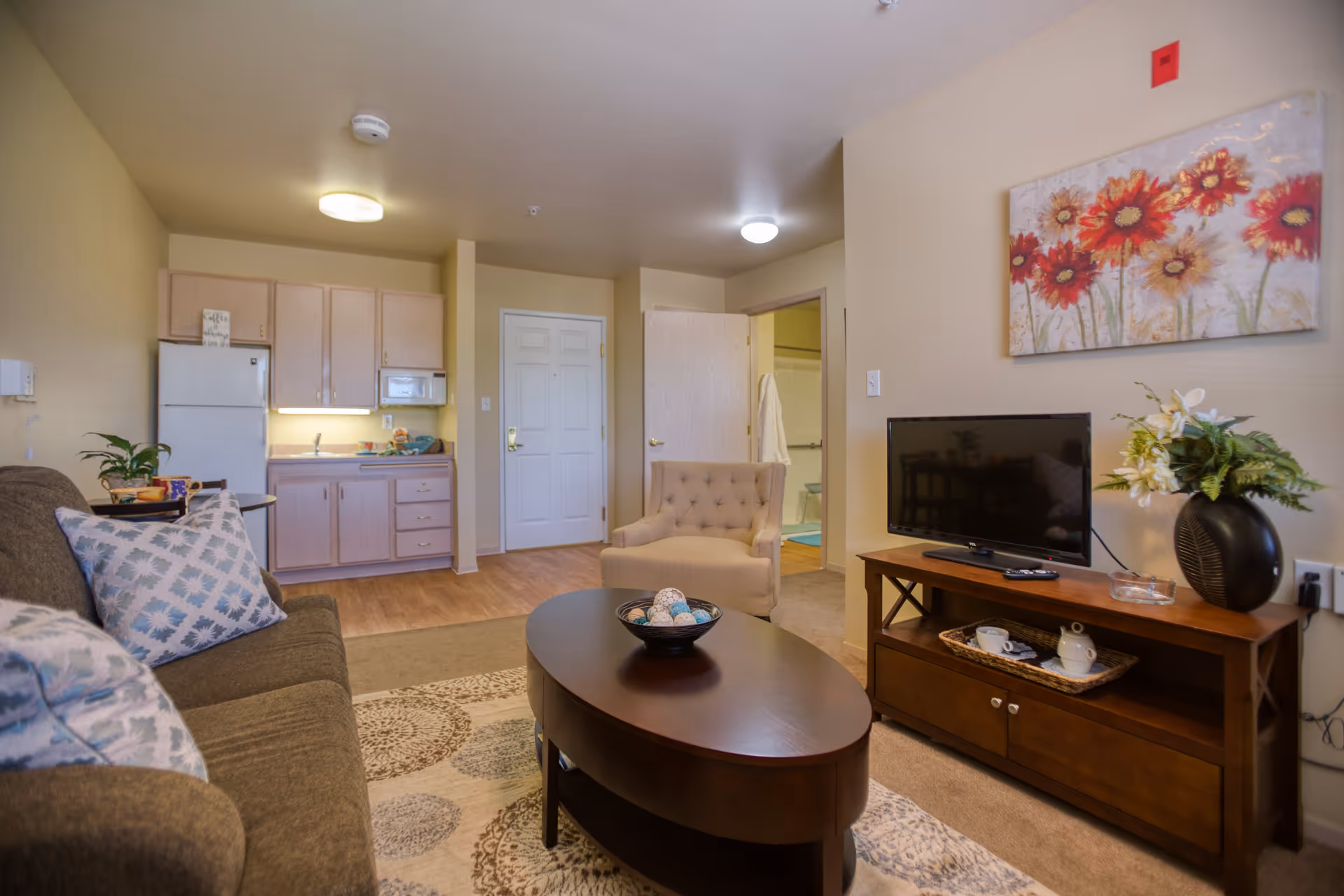 A cozy living area in a senior living facility featuring a brown sofa with patterned pillows, a round wooden coffee table with decorative items, a beige armchair, and a wooden TV stand with a flat-screen TV and a vase of flowers. The background shows a small kitchenette with light wood cabinets, a refrigerator, and a microwave. There is a door leading to a bathroom with a visible toilet and bathrobe hanging on the door. A colorful floral painting hangs on the wall above the TV.
