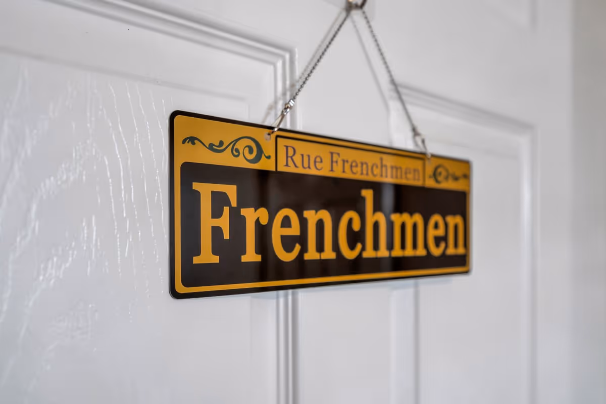 A close-up of a decorative hanging sign on a white door that reads 'Rue Frenchmen Frenchmen' in yellow and black colors.