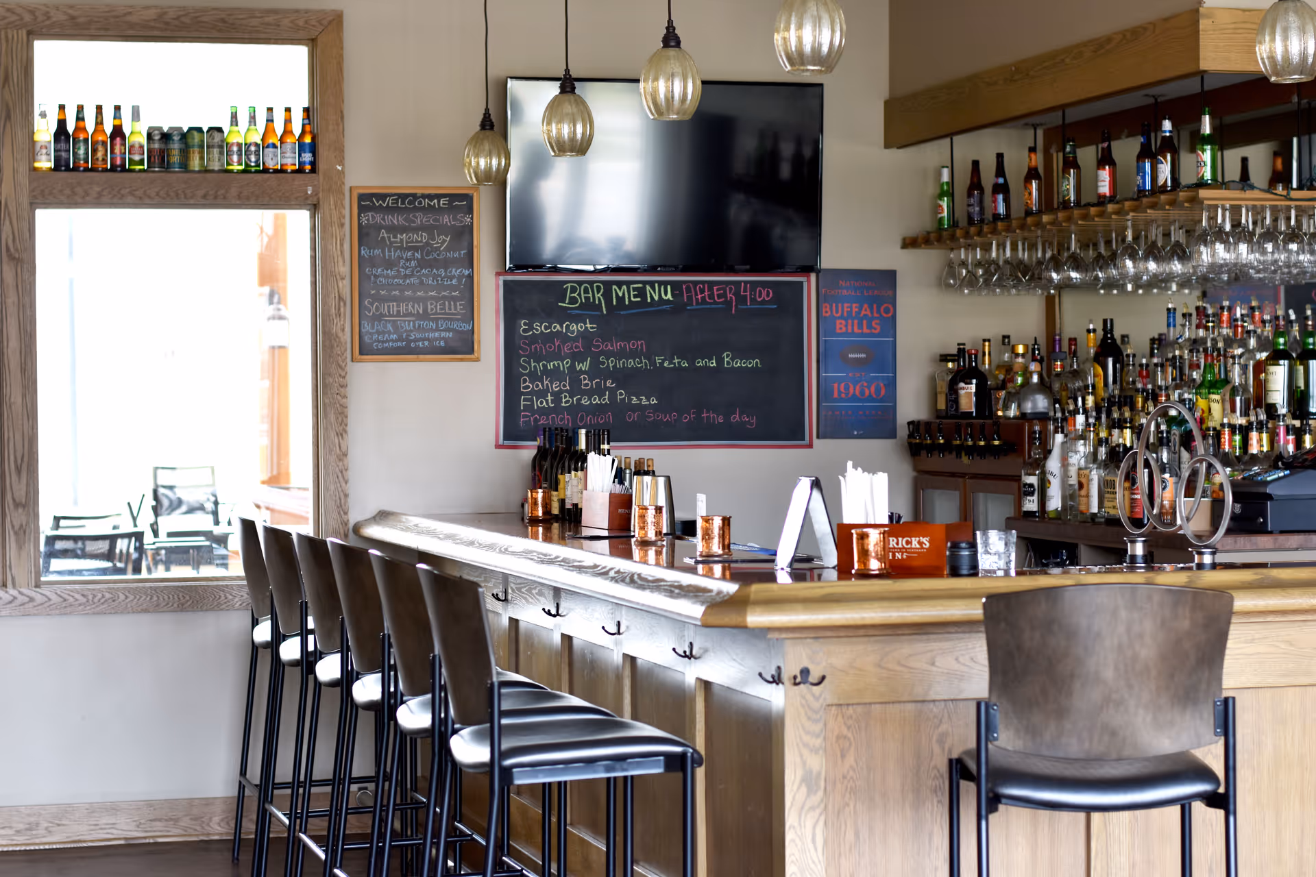 Interior view of a bar area with a wooden counter and several high chairs lined up. Behind the bar, there are shelves stocked with various bottles of liquor and hanging wine glasses. A chalkboard menu and a TV screen are mounted on the wall above the bar. Bottles are displayed on a window ledge to the left, and a small outdoor seating area is visible through the window.
