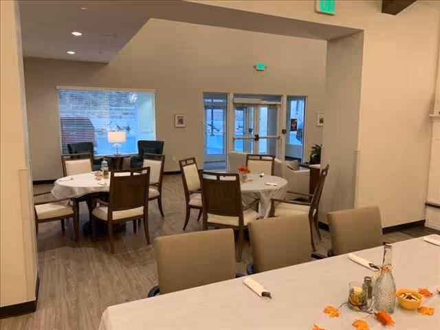 Interior view of a dining area in an assisted living facility with several round tables covered with white tablecloths and surrounded by chairs. The room has light-colored walls, a large window with a lamp on a side table, and a glass door entrance. Some tables have small decorative items on them.