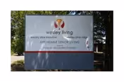 Outdoor sign shows Wesley Living branding and Wesley Pine Meadows near a shaded entrance.