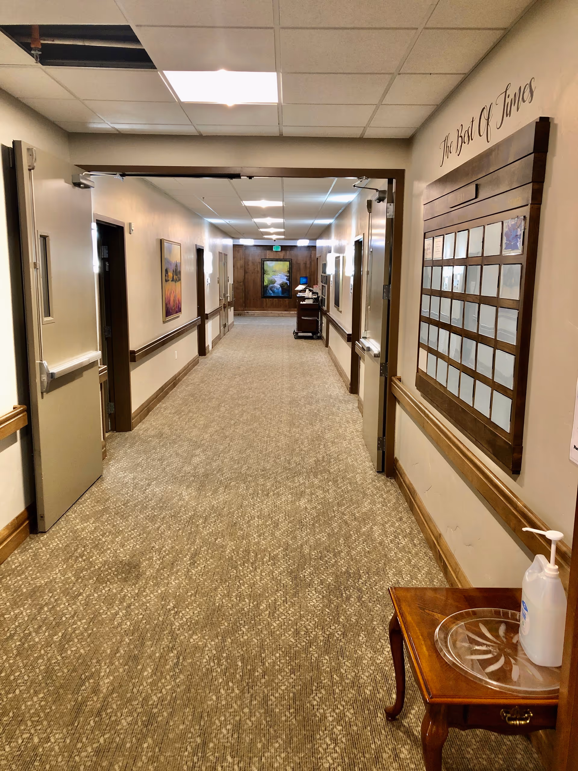 A long, well-lit hallway in an assisted living facility with beige walls and carpeted floor. There are open double doors on the left and right sides, handrails along the walls, framed artwork, and a wooden board with the words 'The Best Of Times' above it on the right wall. A small wooden table with a clear tray and a bottle of hand sanitizer is in the foreground on the right side.