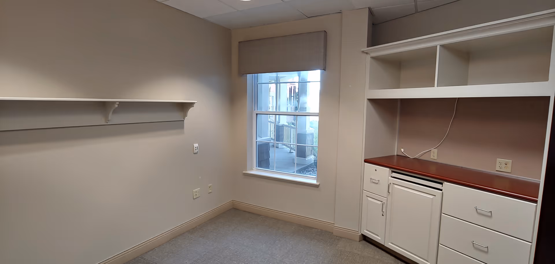 Empty room with beige walls and carpeted floor, featuring a window with a beige valance, a white built-in cabinet with drawers and open shelves, and a long wall-mounted shelf.