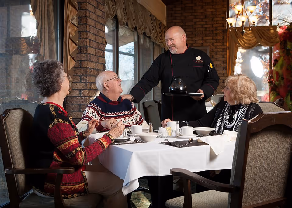 Three elderly people sitting at a dining table with white tablecloth in a warmly lit room, interacting with a smiling chef who is serving coffee from a pot on a tray.