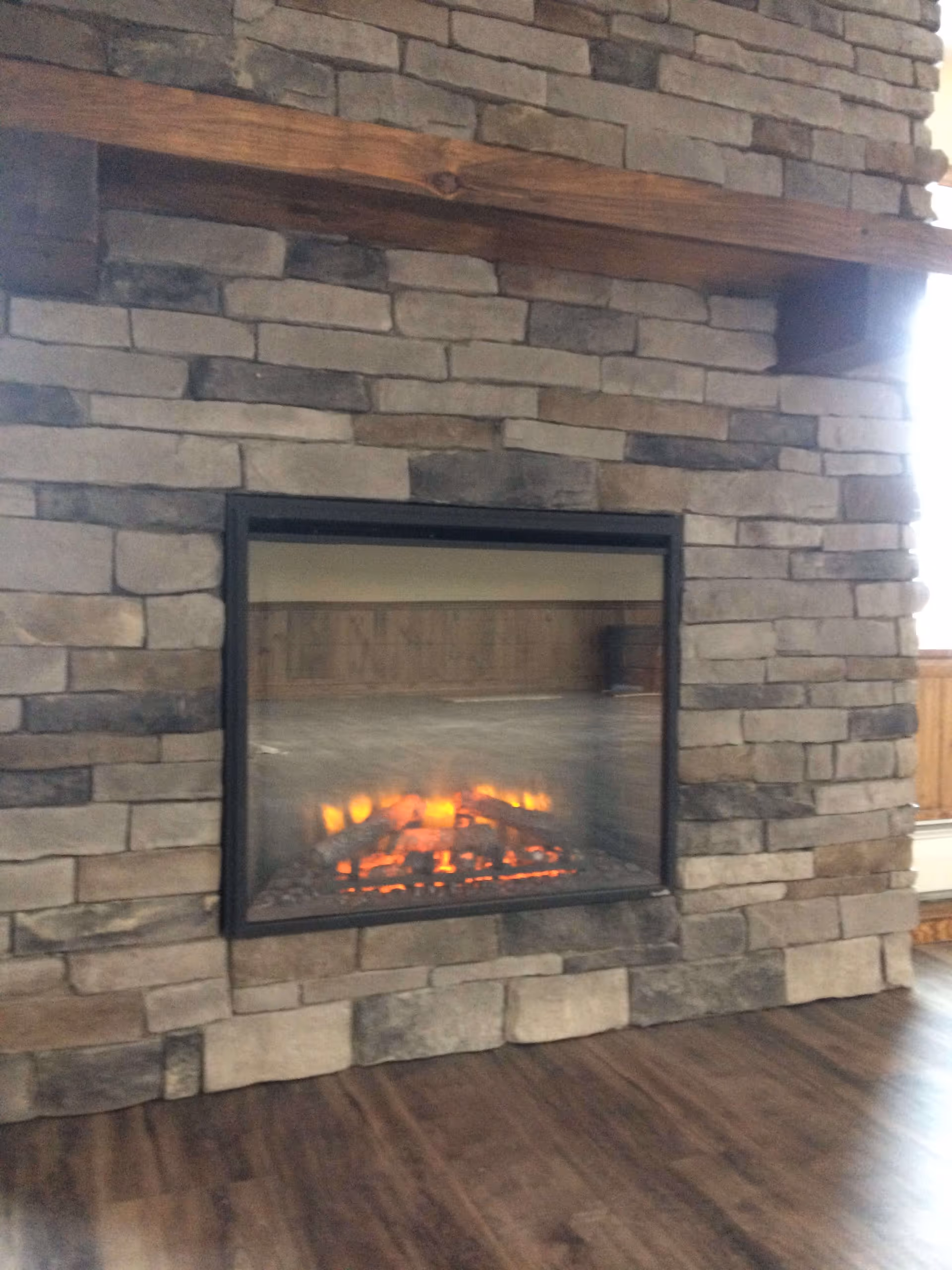 A stone fireplace with a wooden mantel and a glass-enclosed fire burning inside, set against a wall with wood paneling and a wooden floor in front.