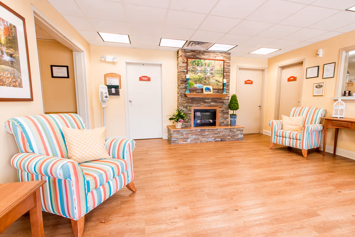 A bright and welcoming interior room with two striped armchairs, each with a decorative pillow, positioned on either side of a stone fireplace. The fireplace has a framed nature painting above it and is flanked by potted plants. The room has light-colored walls, wooden flooring, and several closed doors with red signs. There is a wooden side table and a small desk with framed certificates and a decorative birdcage on it.