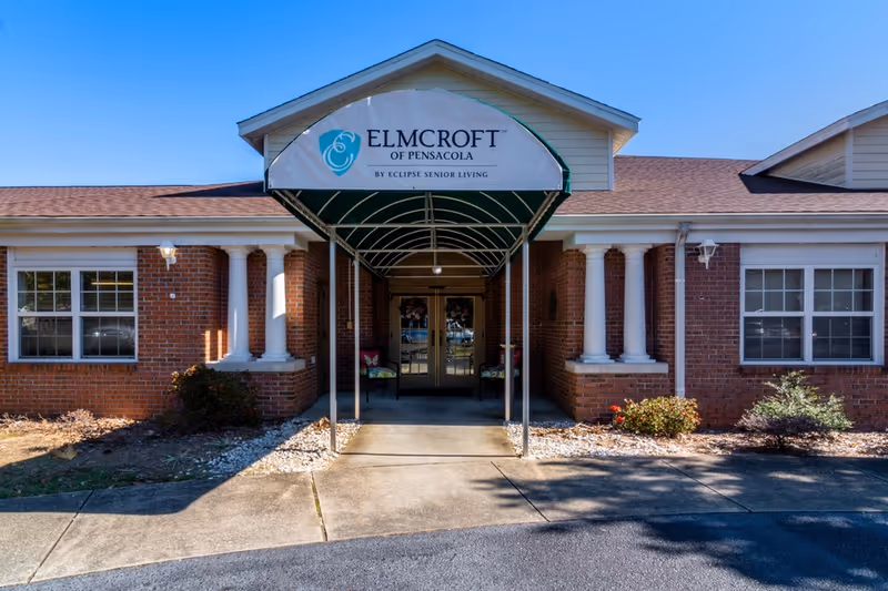 Front entrance of a brick building with white columns and a green awning that reads 'ELMCROFT OF PENSACOLA BY ECLIPSE SENIOR LIVING' under a clear blue sky.