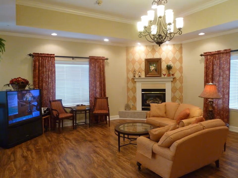 A cozy living room with wooden flooring, two beige sofas with orange cushions, a round glass coffee table, a fireplace with a decorative tiled wall and a framed painting above it, two windows with red patterned curtains, a floor lamp, two wooden chairs with a small table between them, and a television on a stand showing a program.