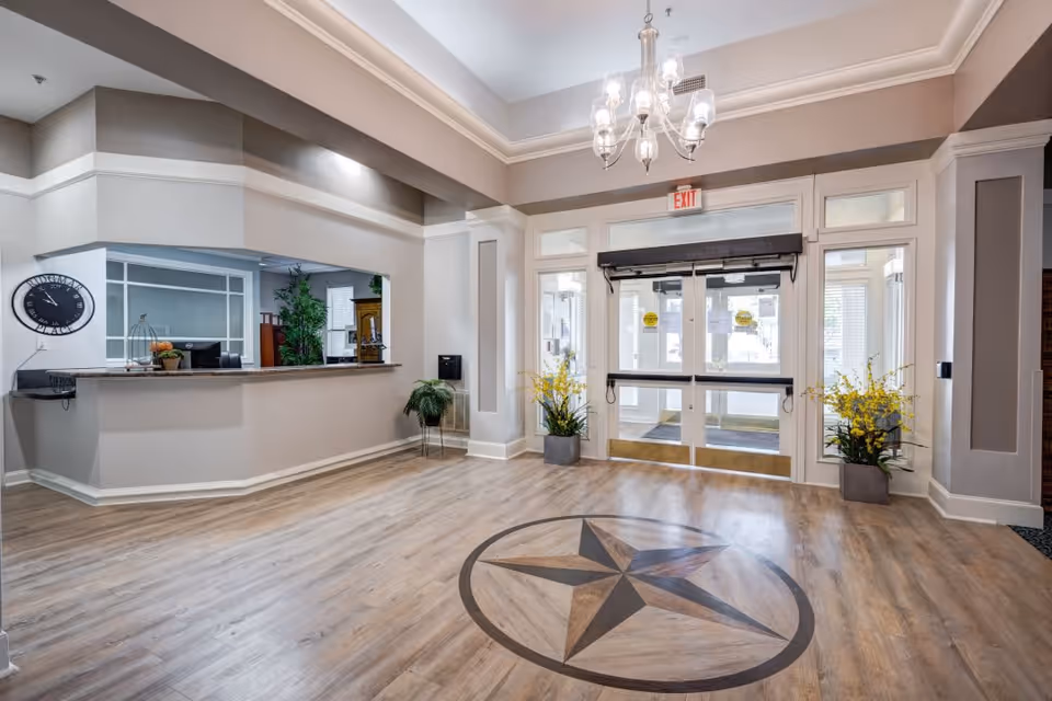 Bright and spacious lobby area of Ridgmar Place with a wooden floor featuring a large star design in the center. There is a reception desk on the left with a clock on the wall above it, and two potted plants with yellow flowers flanking the glass double entrance doors. A chandelier hangs from the ceiling, and the walls are painted in neutral tones.