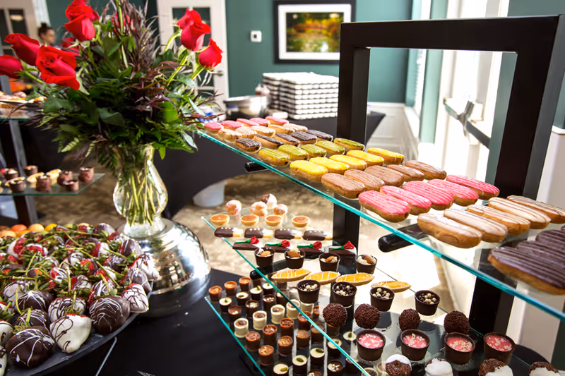 A dessert buffet featuring an assortment of colorful eclairs, chocolate-covered strawberries, and various small pastries arranged on glass shelves. A large vase with red roses and greenery is placed on the table next to the desserts. The background shows a green wall with a framed picture and a window letting in natural light.