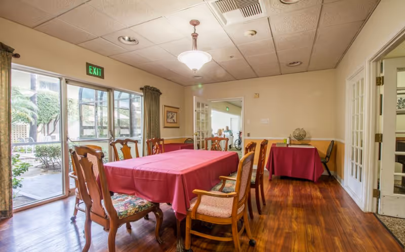 Dining room with a long table covered in a red tablecloth surrounded by wooden chairs, hardwood floors, and glass doors leading to an outdoor courtyard.