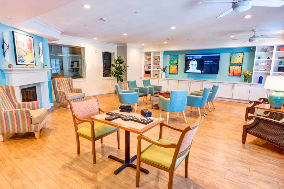 A bright and spacious living room area in a senior living facility with wooden flooring, multiple seating options including striped armchairs, teal upholstered chairs, and wooden chairs with green cushions. A table in the foreground has a large domino set on it. The room features a white fireplace with colorful artwork above it, built-in white cabinets with decorative items, a large flat-screen TV mounted on a teal accent wall, ceiling fans, and a table lamp on a side table.