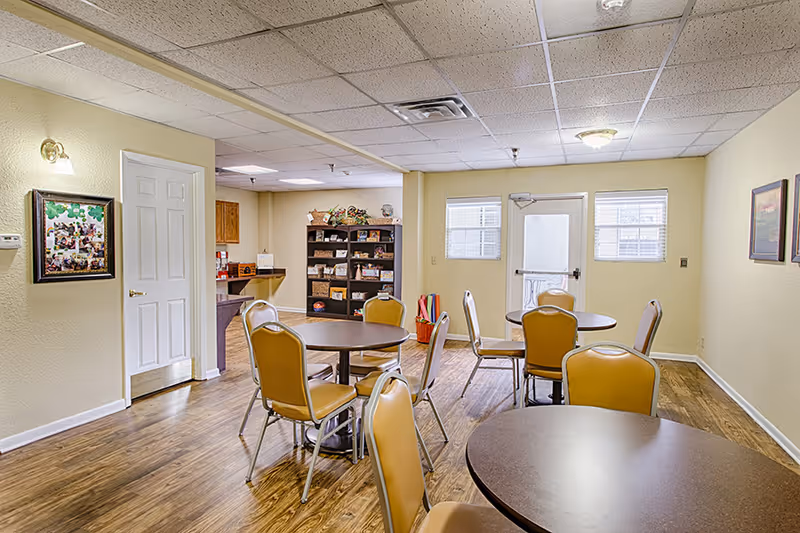 A communal dining/activity room with round tables, yellow chairs, shelving and a rear door.