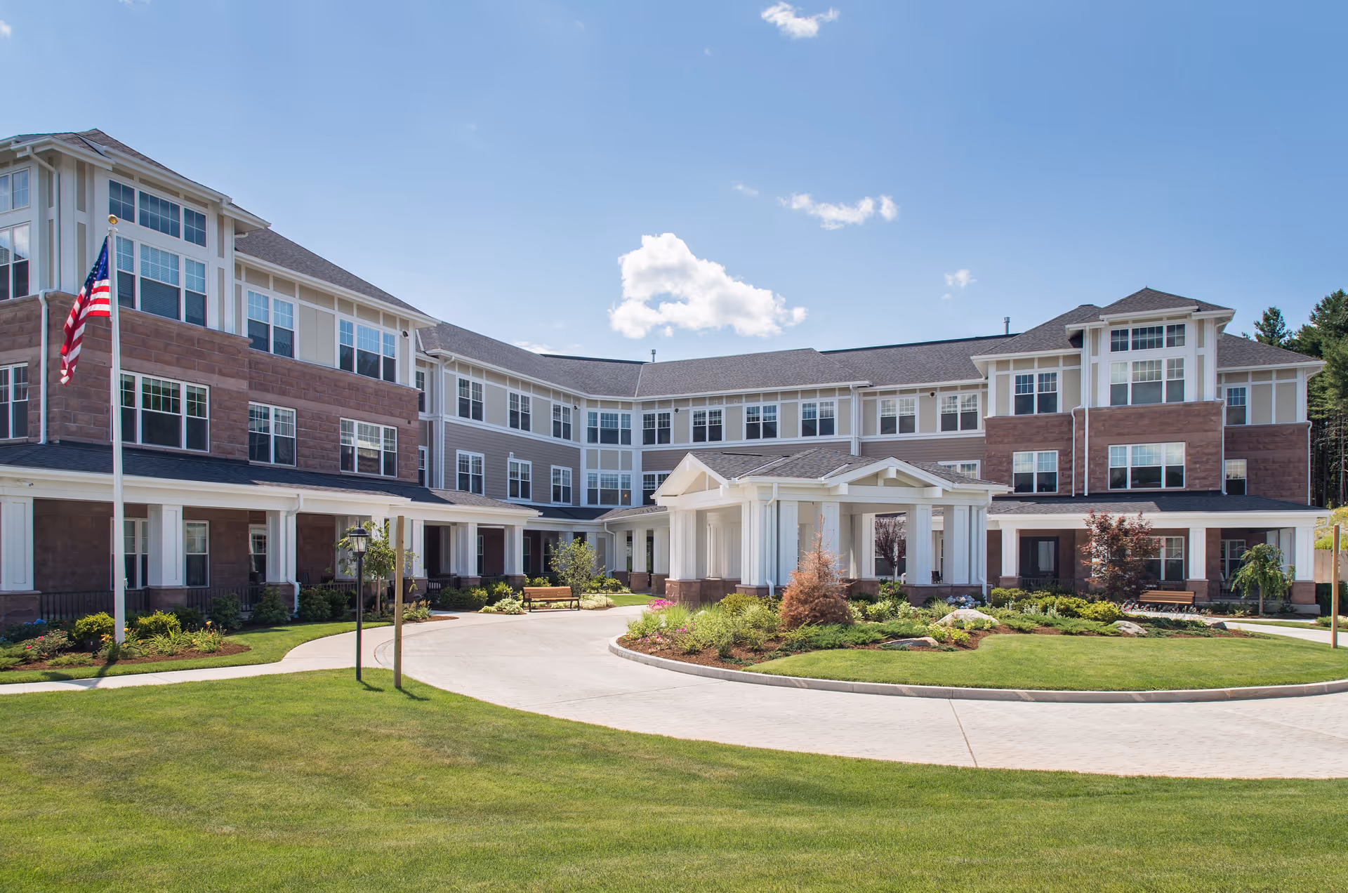 Exterior view of a large, modern senior living facility building with three stories, featuring a covered entrance, landscaped gardens, benches, and an American flag on a flagpole under a blue sky with some clouds.