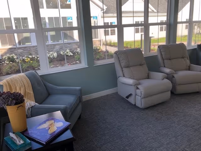 A cozy sitting area with two beige recliner chairs and a blue armchair with a yellow blanket draped over it. A small black side table holds a potted plant, a box of tissues, and a photo album. Large windows let in natural light and show a view of a garden and neighboring buildings outside.