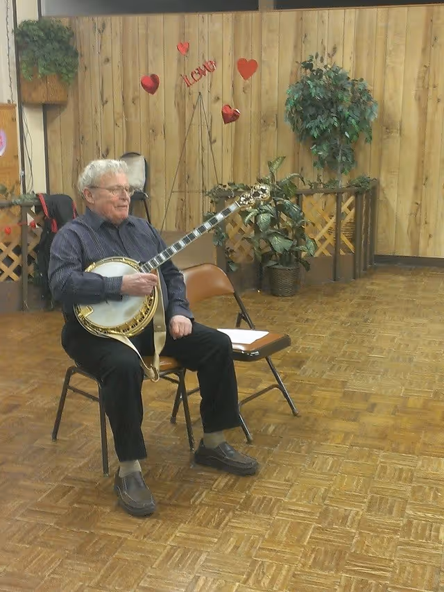 An elderly man sitting on a chair playing a banjo in a room with wooden panel walls and parquet flooring. The room is decorated with potted plants and red heart decorations on the wall, including a sign that says 'I LOVE YOU'. Another empty chair with a piece of paper on it is next to him.