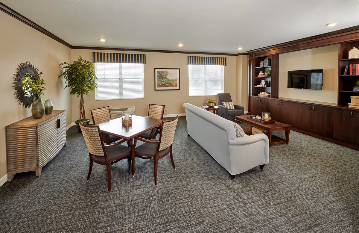 A well-lit living room with a gray couch, a wooden coffee table, a single armchair, and a built-in dark wood entertainment center with shelves and a mounted TV. There is a dining table with four chairs in the foreground, a sideboard with decorative vases and a mirror on the left, and two windows with striped valances letting in natural light.