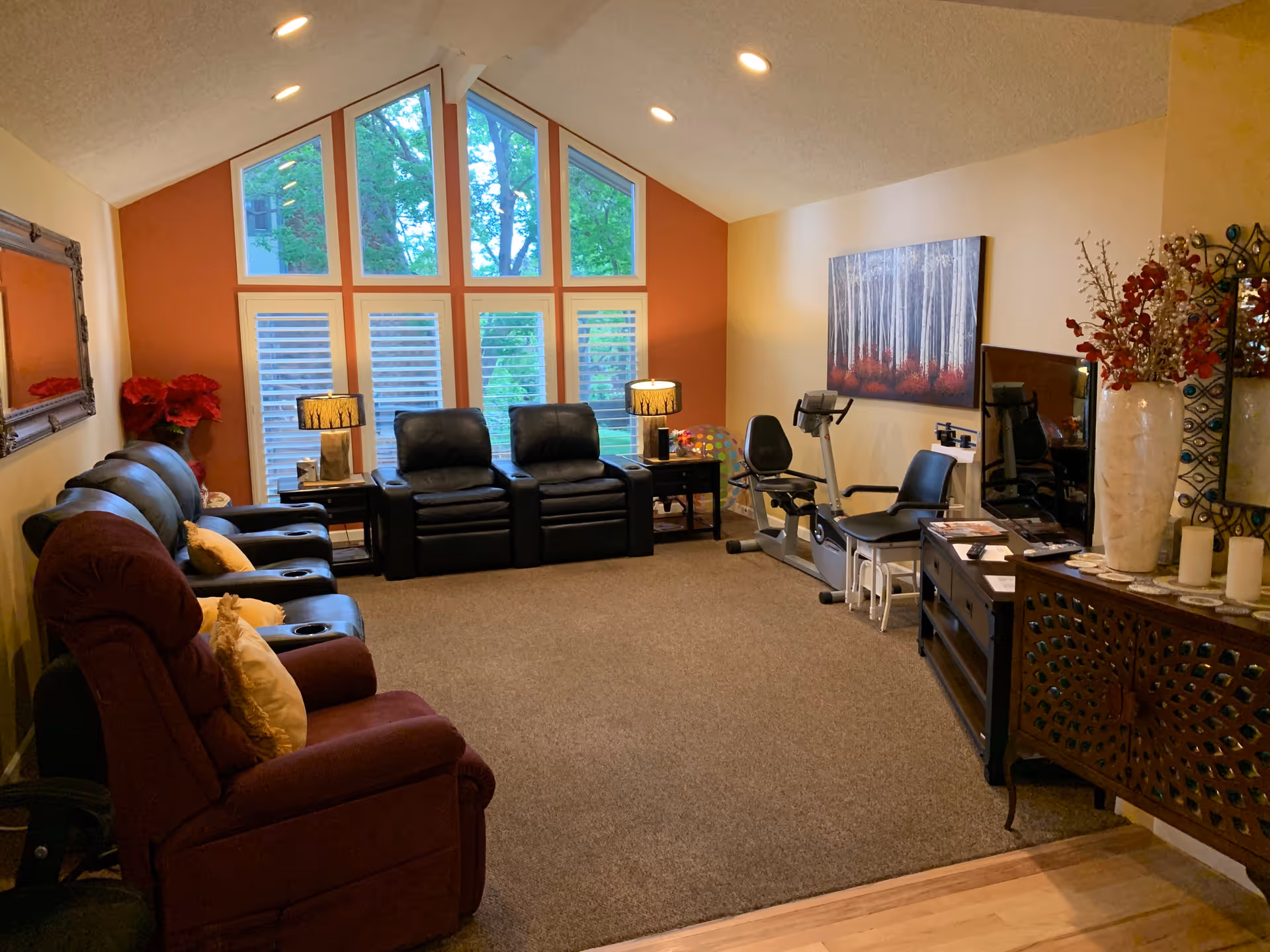 A cozy living room with a row of recliner chairs along the left wall and two black recliners facing large windows with a view of trees. The room has warm lighting from table lamps, a painting of birch trees on the right wall, exercise equipment, and decorative vases with flowers on a wooden cabinet.