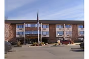 Exterior view of a three-story brick and beige panel building with multiple windows, an entrance with a covered awning, and an American flag flying on a flagpole in front. Several cars are parked in the parking lot in front of the building under a clear blue sky.