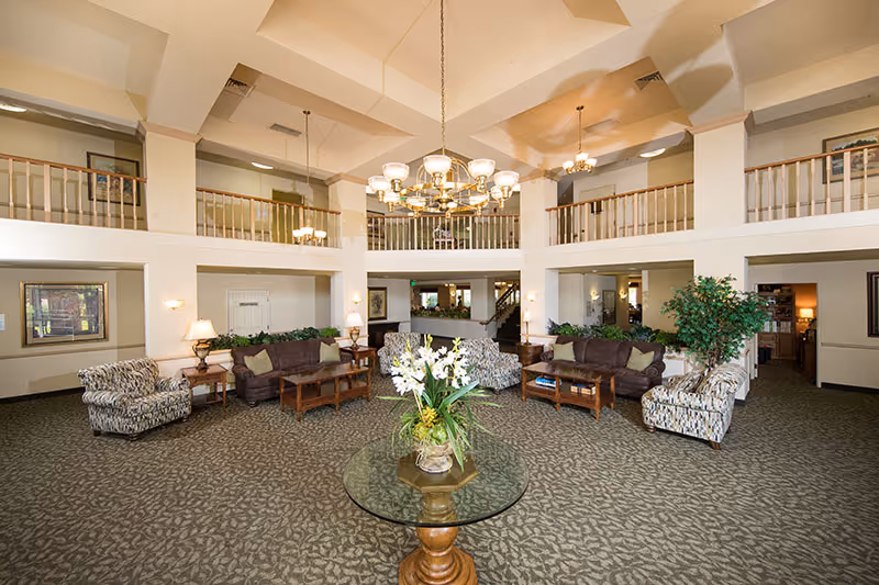Spacious and well-lit living room area in Bethel Retirement Community featuring multiple seating arrangements with patterned and solid sofas and armchairs, a round glass table with a floral centerpiece in the foreground, carpeted floor, and a large chandelier hanging from a high ceiling with a second-floor balcony overlooking the space.