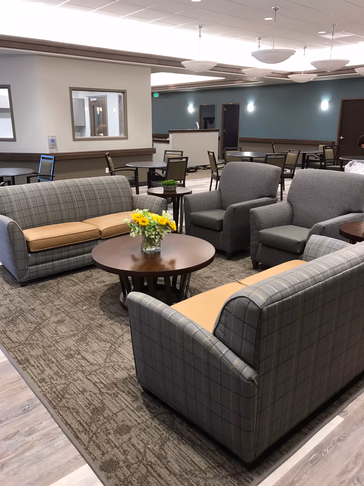 A spacious senior living common lounge with sofas, armchairs, round coffee tables and a dining area in the background.
