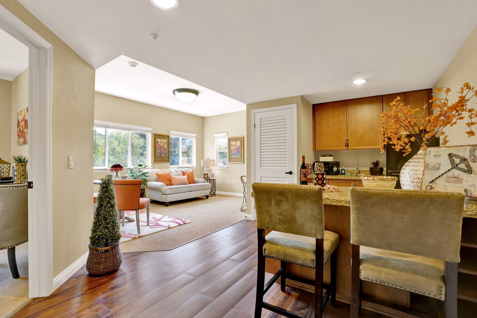 Interior view of a senior living facility showing a cozy living room with a white sofa, orange cushions, and two framed pictures on the wall. Adjacent to the living room is a kitchen area with wooden cabinets, a granite countertop, two upholstered bar stools, and decorative items including a vase with orange flowers. The floor is a combination of wood and carpet, and there is natural light coming through the windows.
