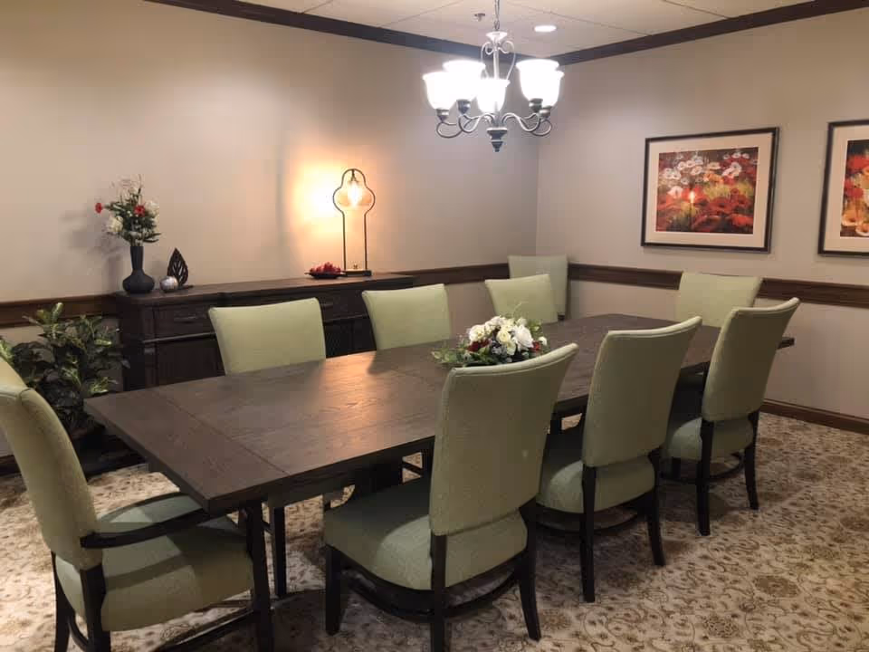 A dining room with a long dark wooden table surrounded by eight light green upholstered chairs. A floral centerpiece is on the table. The room has beige walls with two framed floral paintings, a chandelier hanging from the ceiling, a sideboard with decorative items, and a floor lamp emitting warm light. There are also some potted plants in the corner.