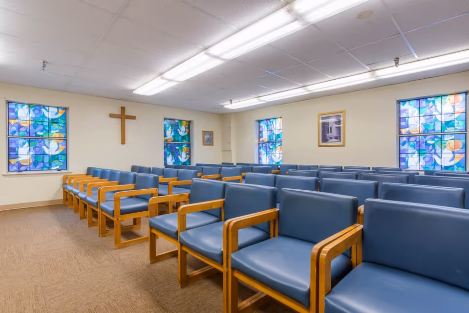 Interior view of a chapel or small worship room with rows of blue cushioned wooden chairs, stained glass windows featuring colorful abstract designs, a wooden cross mounted on the wall, and framed pictures on the walls.
