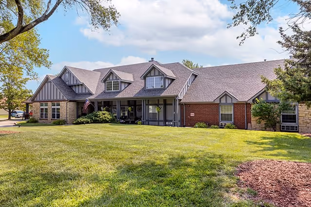 Exterior view of a single-story residential building with a combination of brick and stone walls, multiple gabled roofs, and a covered porch. The building is surrounded by a well-maintained lawn with trees and shrubs under a partly cloudy sky.