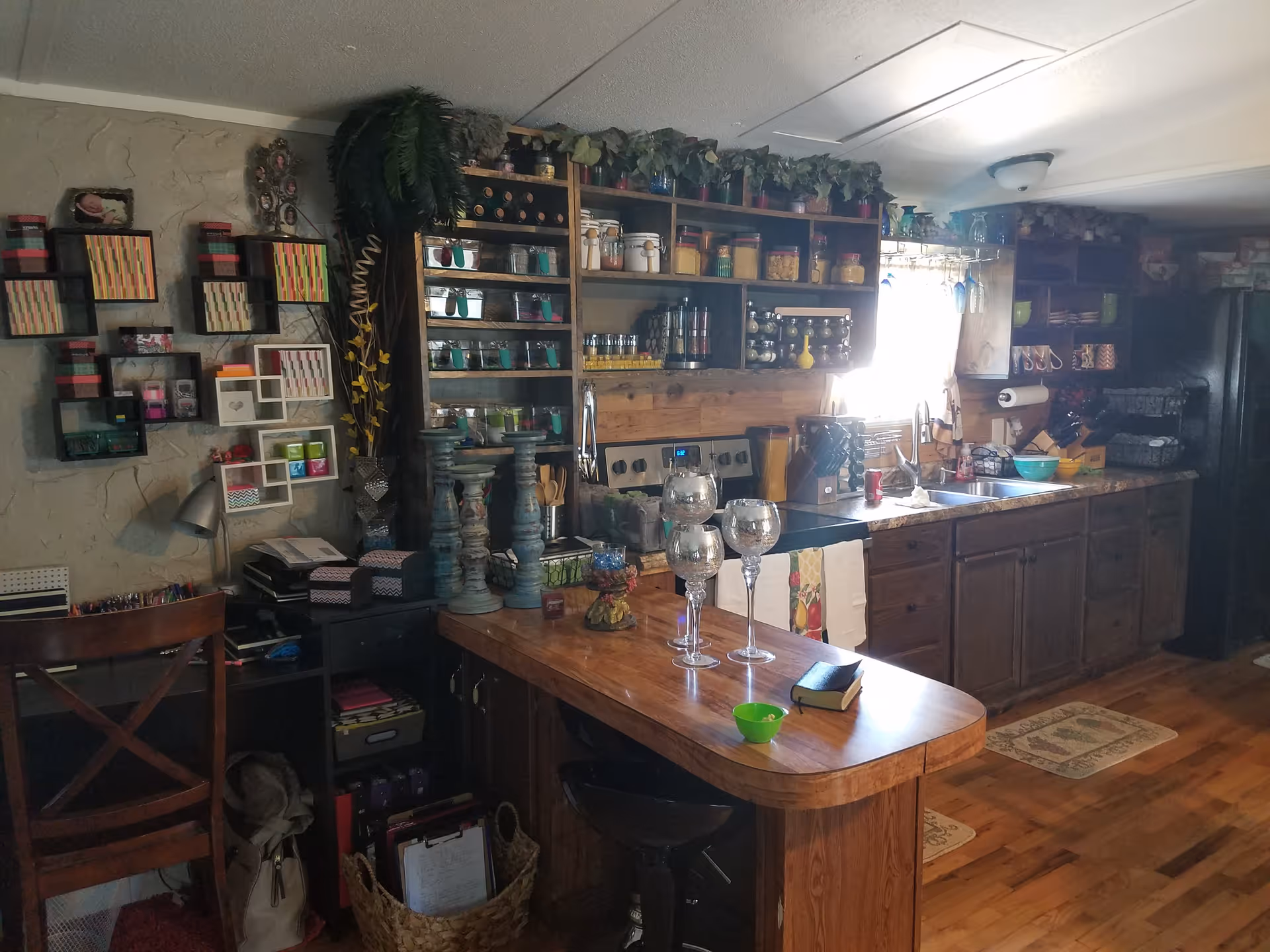 A cozy kitchen area with wooden cabinets and countertops. The kitchen features a stove, sink, and various jars and containers on open shelves above the counter. There are decorative plants on top of the shelves and several large candle holders on the counter. A small table or island with three large wine glasses, a small green bowl, and a book is in the foreground. The walls have decorative shelving with colorful boxes and other items. The floor is wooden with small rugs near the sink area.