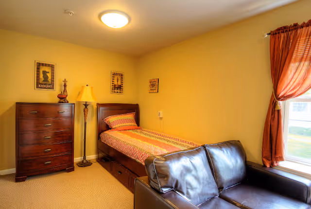 Small furnished bedroom with a single bed, wooden dresser, floor lamp and leather couch by a curtained window.