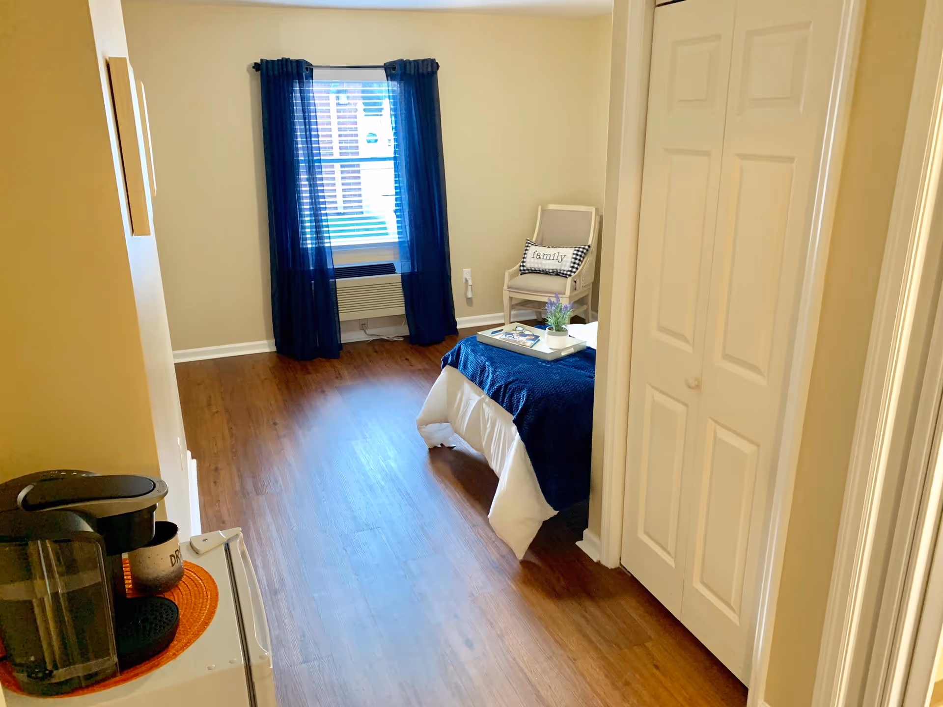 Furnished bedroom with wood floors, a single bed topped with a navy throw, a chair by a window with dark curtains, and a coffee maker on a small table near the door.