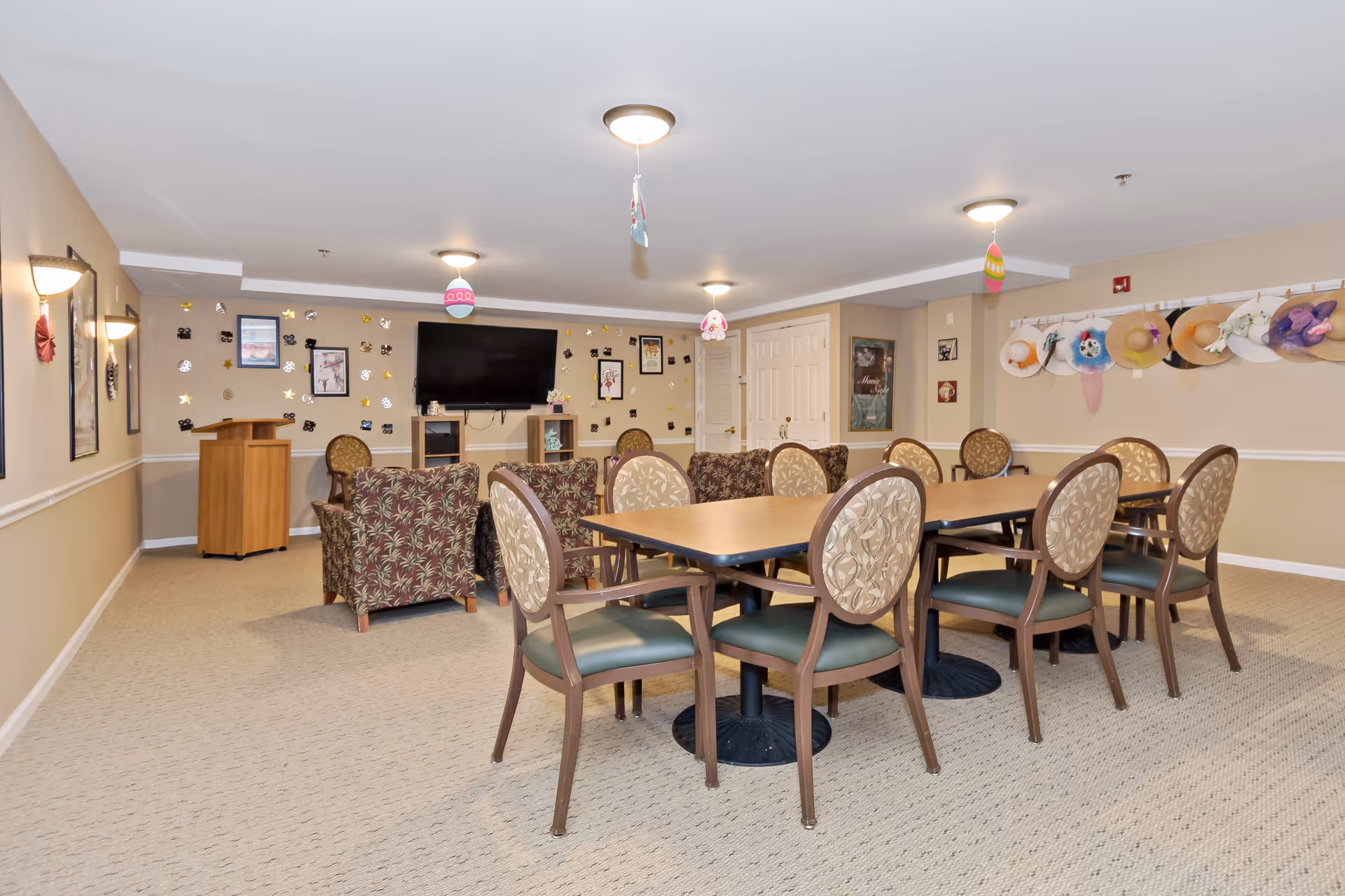 A communal room in an assisted living facility featuring a long table with eight chairs around it. The room has beige walls decorated with hanging hats on one side and various framed pictures and decorations on the far wall. There are two patterned armchairs facing a wall-mounted flat-screen TV. The ceiling has several light fixtures and hanging decorative ornaments.