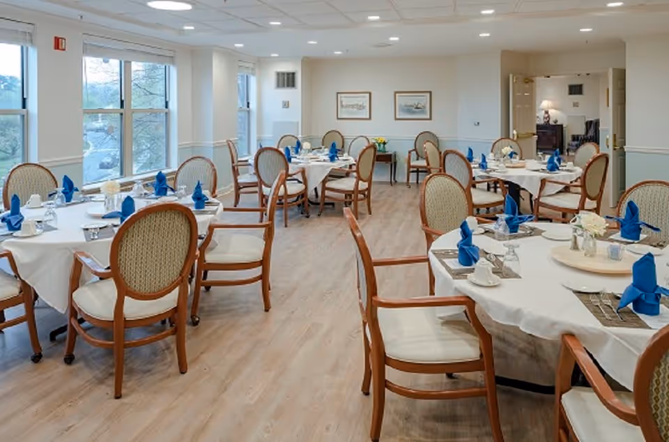 A bright dining room with multiple round tables covered with white tablecloths, each set with blue folded napkins, cups, and glasses. The room has large windows letting in natural light, wooden chairs with cushioned seats and backs, light wood flooring, and framed artwork on the walls.
