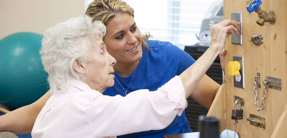 An elderly woman and a younger woman are interacting with a wooden activity board that has various switches, locks, and knobs. The elderly woman is reaching to flip a switch while the younger woman, wearing a blue shirt, smiles and supports her. A large green exercise ball is visible in the background.