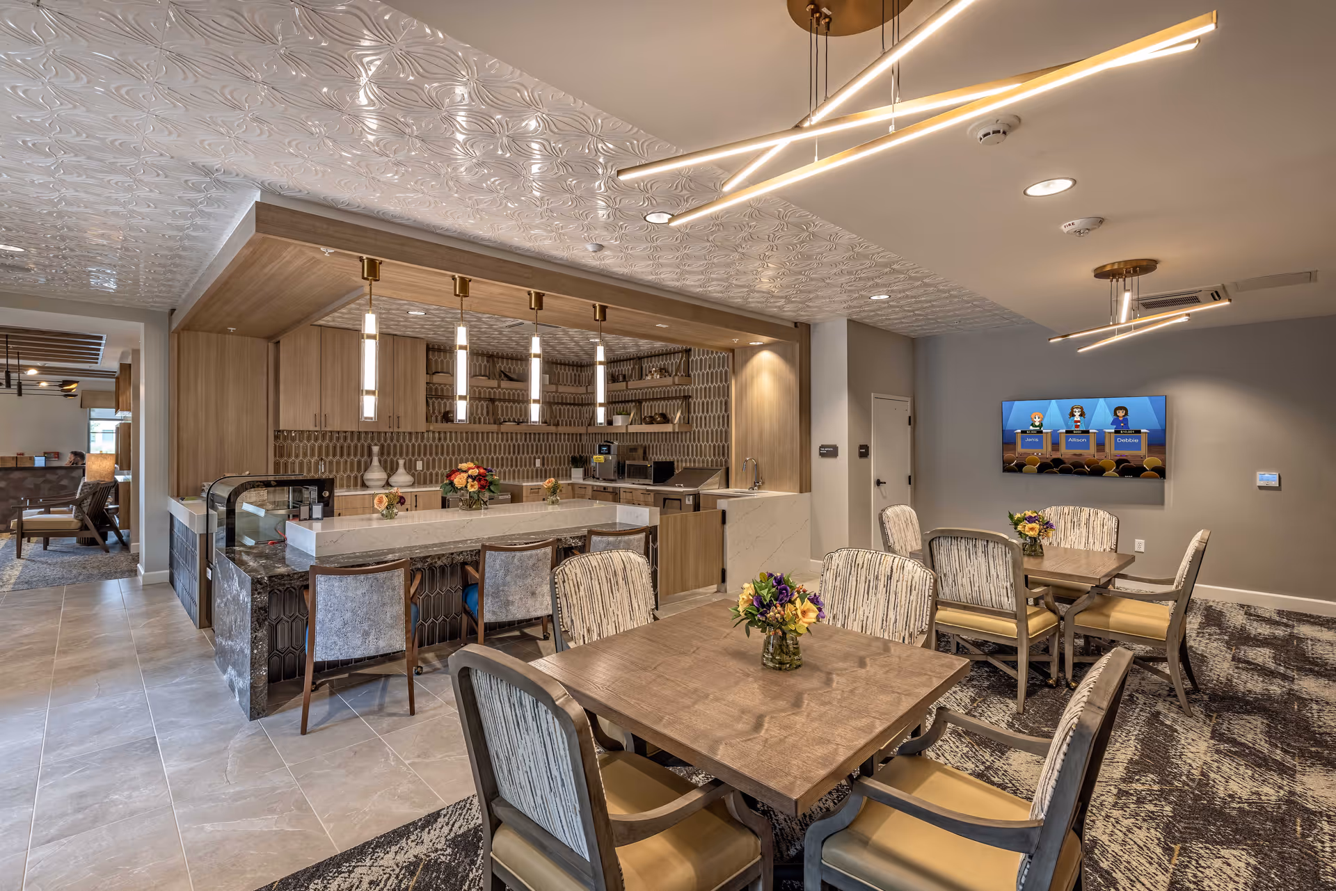 A modern dining area in a senior living facility featuring two square wooden tables with six cushioned chairs each. The tables have small floral centerpieces. Behind the dining area is a kitchen bar with four high chairs, pendant lights, and a tiled backsplash. A flat-screen TV is mounted on the wall displaying a cartoon quiz show. The ceiling has decorative tiles and contemporary light fixtures.