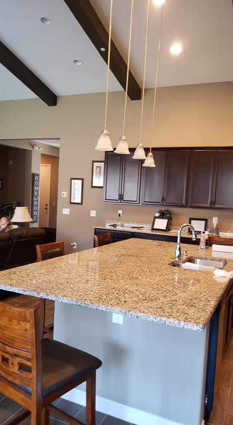 Interior view of a kitchen area with a large granite countertop island featuring a built-in sink and faucet. Four pendant lights hang above the island. Dark wooden cabinets are mounted on the wall in the background. Wooden chairs are placed around the island. Part of a living room with a person sitting on a couch is visible to the left.