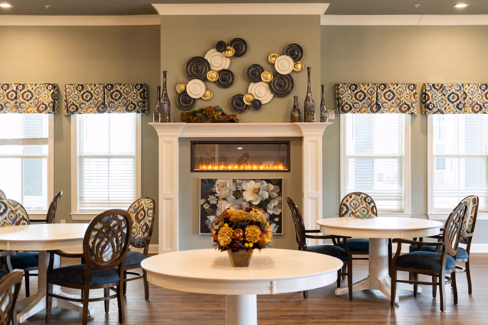 A cozy dining area with round white tables and wooden chairs with patterned cushions. The room features a modern electric fireplace with decorative plates and vases on the mantel. There are four windows with patterned valances letting in natural light, and a floral centerpiece on the table in the foreground.