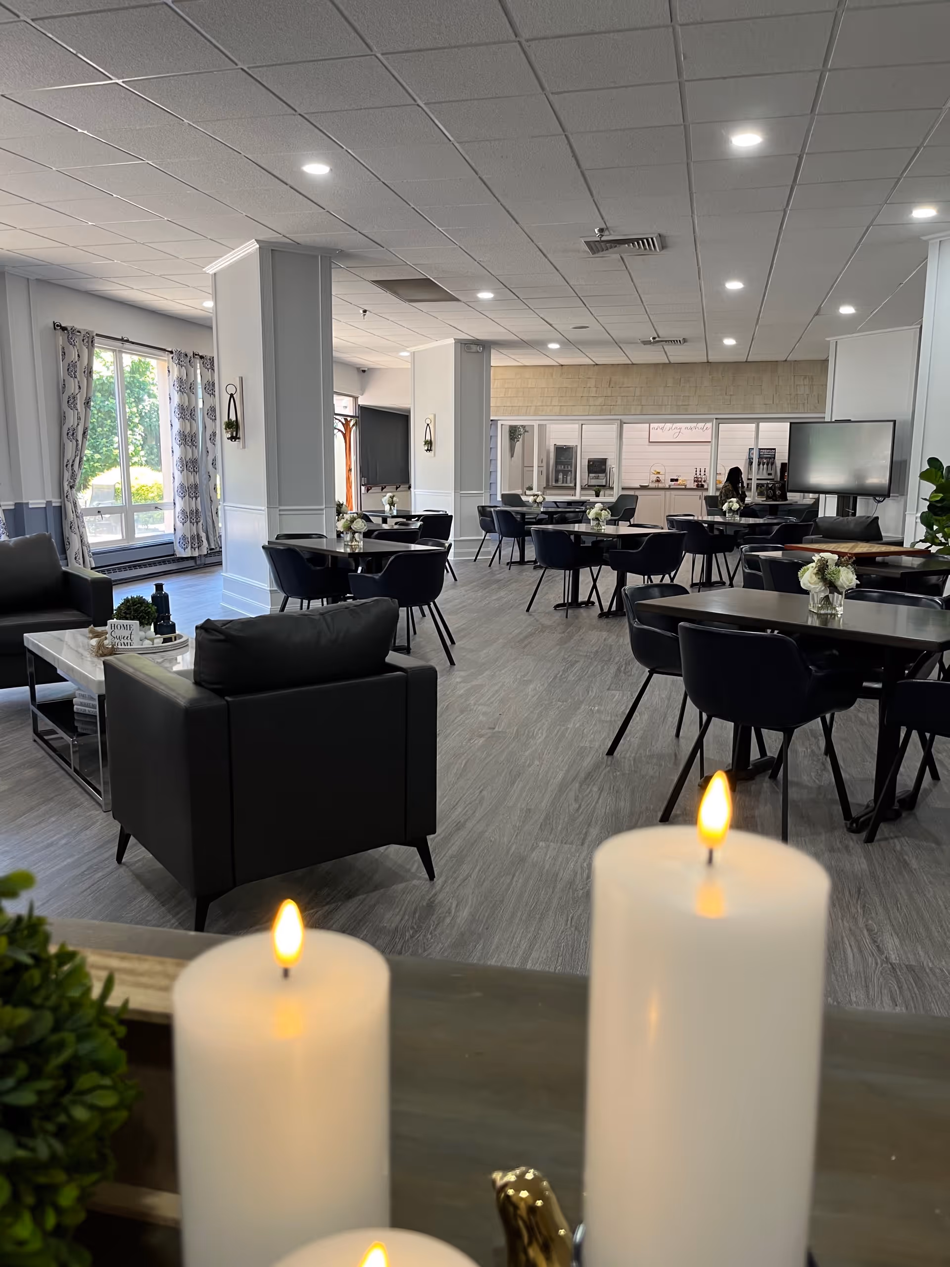 A spacious, well-lit common area with multiple black chairs and tables arranged neatly. There are large windows with patterned curtains allowing natural light to enter. The room features modern decor with white walls, a gray floor, and ceiling lights. In the foreground, there are three lit white candles and some greenery on a table.