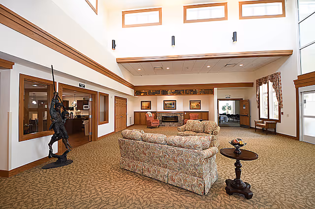 A spacious and well-lit living room area with high ceilings and multiple windows near the ceiling allowing natural light. The room features a patterned sofa and loveseat set arranged around a small round wooden table with a decorative bowl. There is a fireplace on the far wall with framed artwork above it and two armchairs on either side. To the left, there is a statue of a knight holding a spear near a doorway leading to another room. The room has beige carpet with a subtle pattern and wood trim accents.