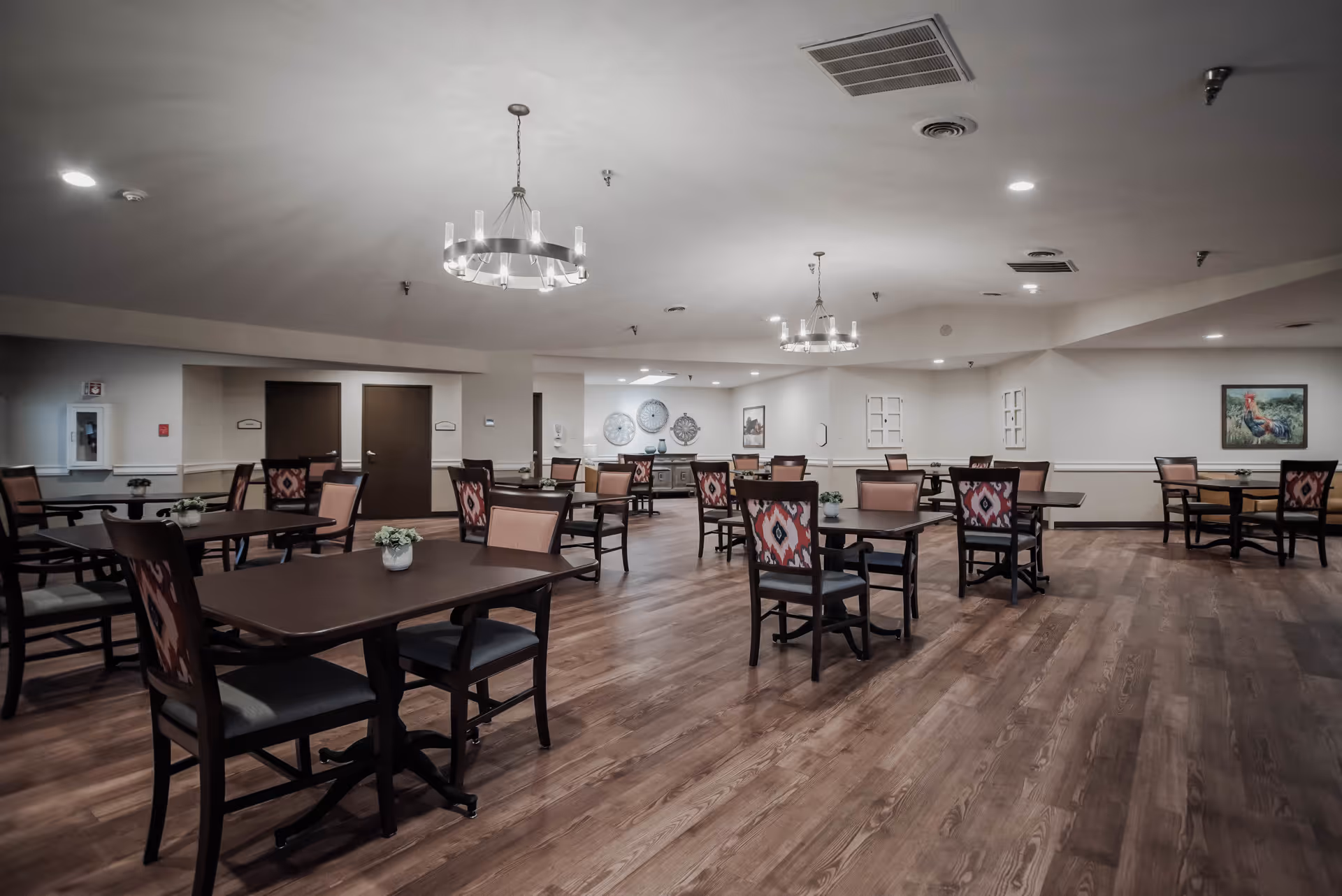 A spacious dining room with multiple dark wood tables and chairs arranged neatly. The chairs have patterned upholstery on the backrests. The room has wooden flooring, light-colored walls, two modern chandeliers hanging from the ceiling, and decorative wall art. There are no people present.
